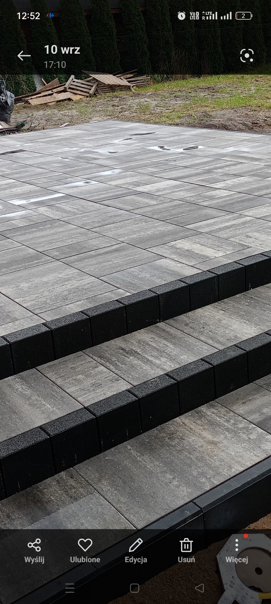 Ułożona nawierzchnia z szarych kostek brukowych z czarnym obrzeżem, widoczne schody z kostki, w tle trawa i drzewa.