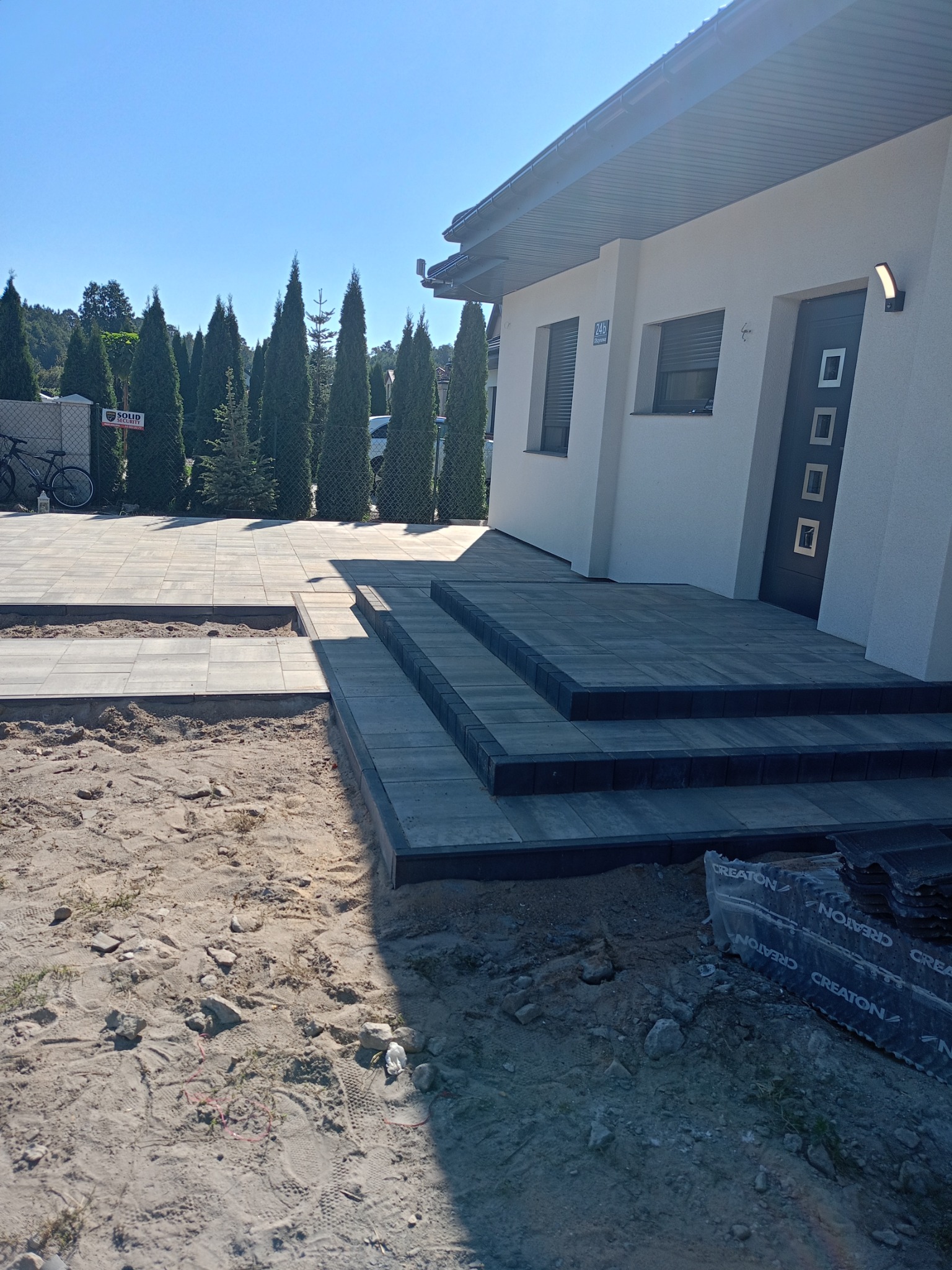 Nowo ułożony taras z szarej kostki brukowej z trzema stopniami prowadzącymi do drzwi wejściowych domu, widoczne pakiety kostki brukowej Creaton, w tle rząd wysokich tuj.