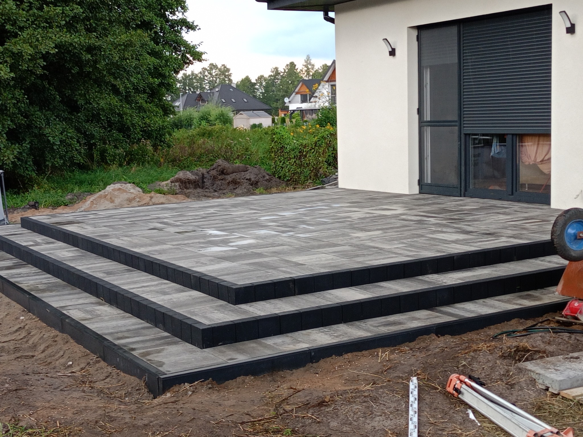 Nowo ułożony taras z kostki brukowej z trzema stopniami, obrzeża z ciemnych kostek, widok na dom z zamkniętymi roletami i ogród w tle, teren wokół tarasu jeszcze niezagospodarowany.