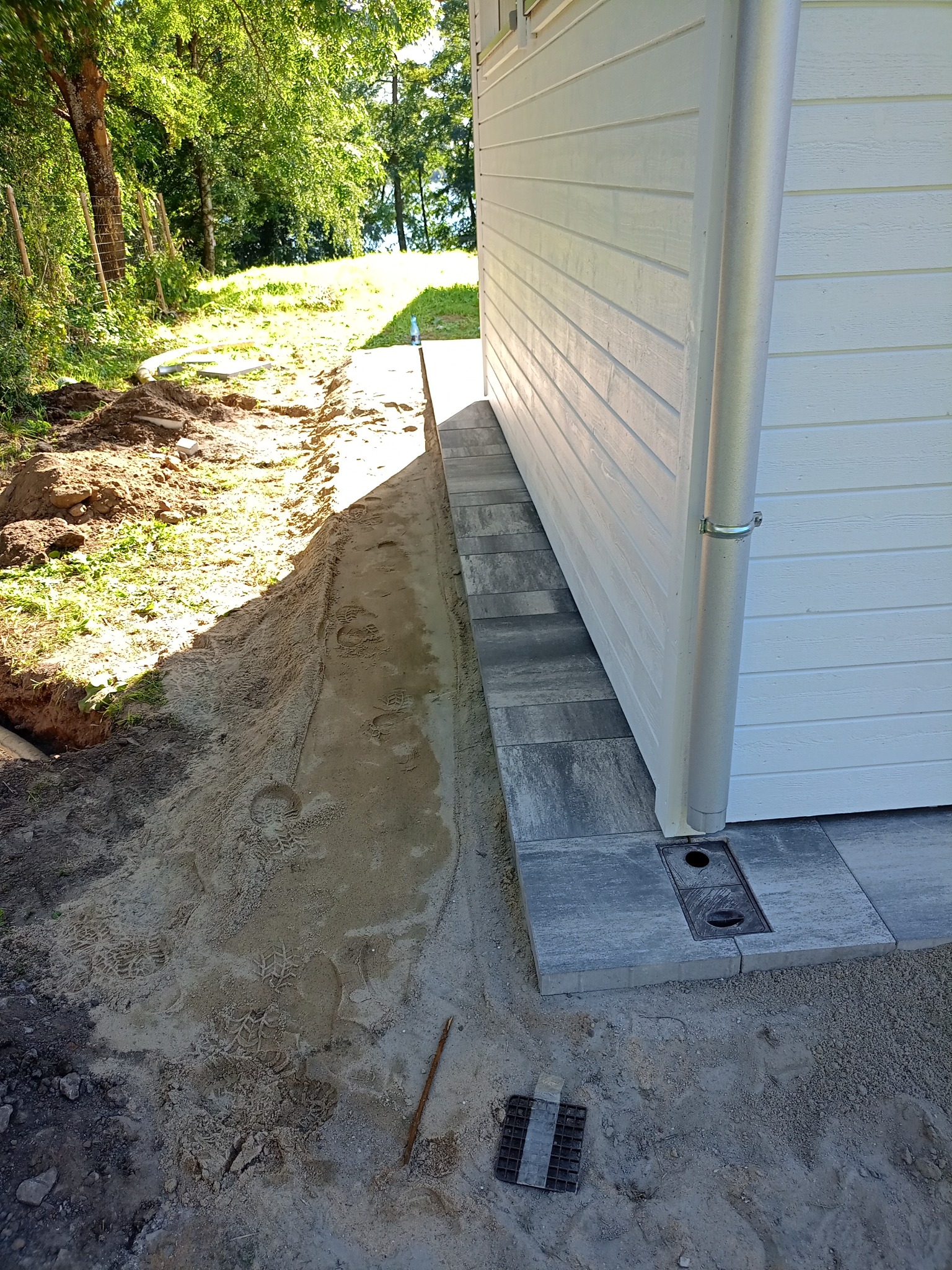 Przygotowanie podłoża pod ścieżkę z kostki brukowej obok białego budynku, widoczne ślady stóp w piasku i studzienka odpływowa.