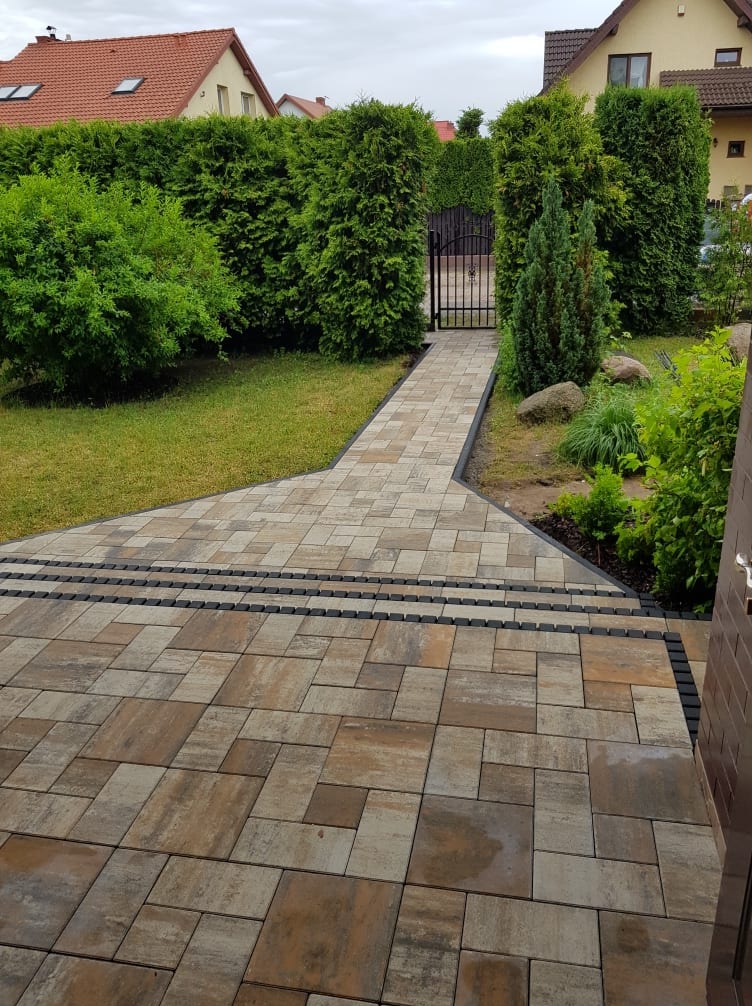 Ułożona kostka brukowa w odcieniach brązu i beżu prowadząca do furtki w żywopłocie z tui, z czarnymi obrzeżami i pasami z kostki.