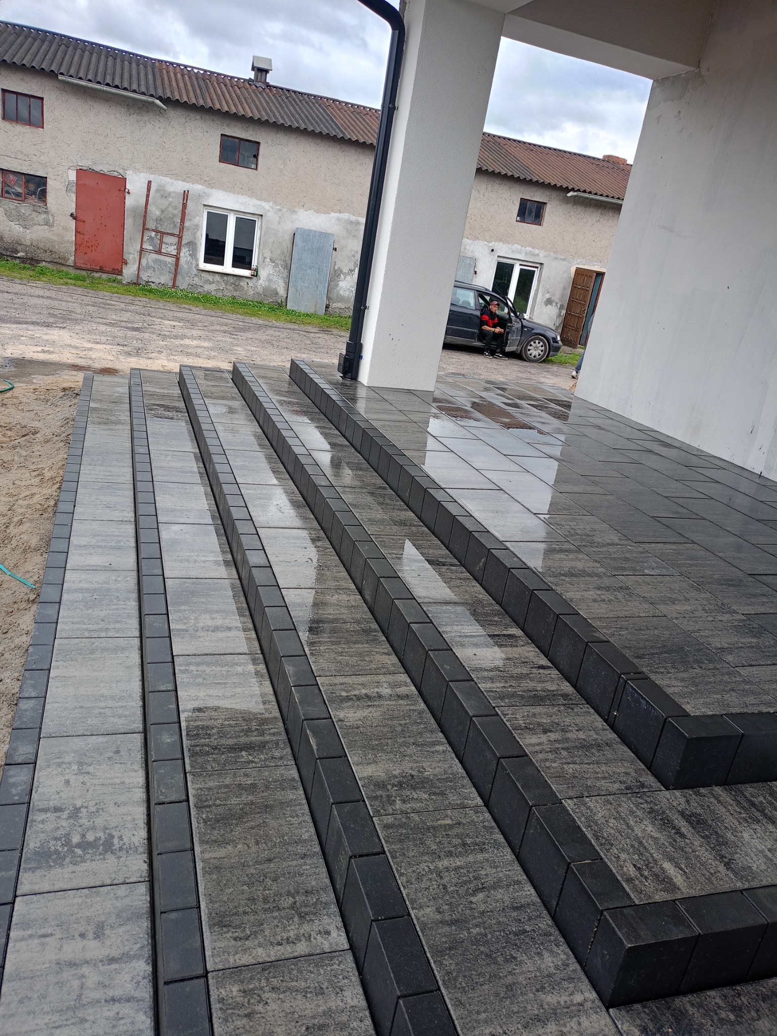 Ułożona, mokra kostka brukowa w odcieniach szarości prowadząca do wejścia budynku z widocznym samochodem w tle.
