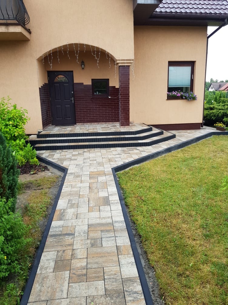 Ułożona ścieżka z kostki brukowej prowadząca do wejścia domu z trzema schodami, obrzeża w kolorze grafitowym, trawnik po prawej stronie, kwiaty w doniczce na parapecie okna.