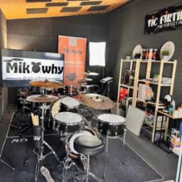 Studio perkusyjne z zestawem perkusyjnym, wygłuszonymi ścianami, ekranem z logo szkoły perkusyjnej MikWhy Drum School i regałem z akcesoriami perkusyjnymi.