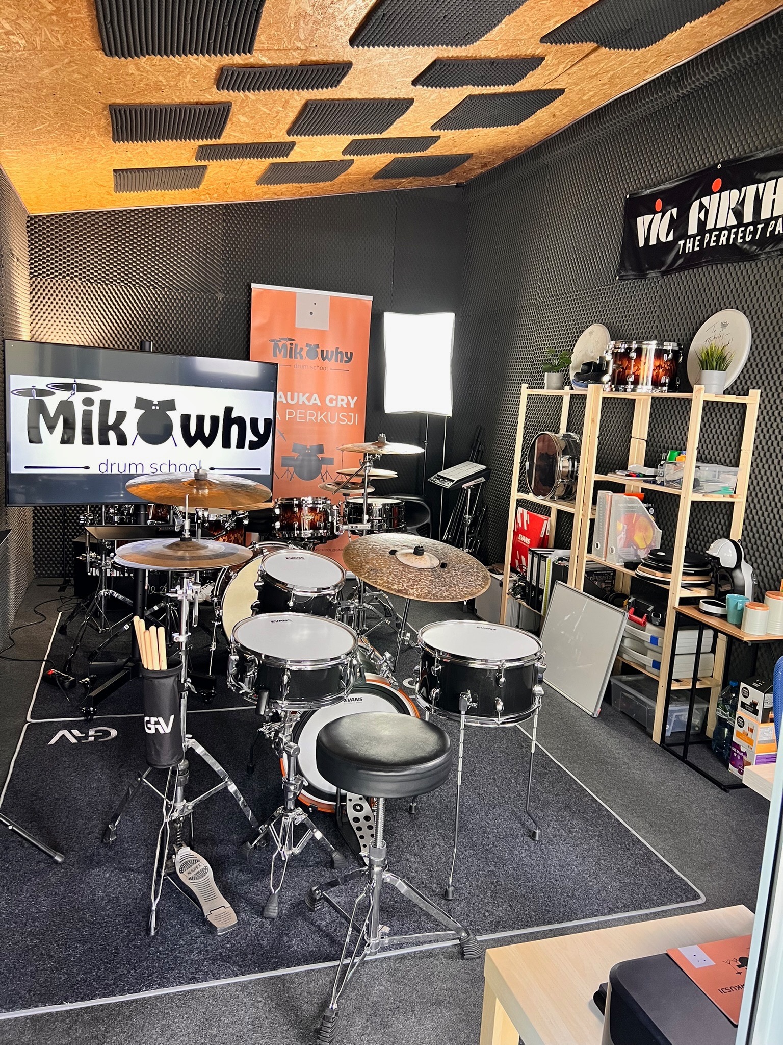 Studio perkusyjne z zestawem perkusyjnym, wygłuszonymi ścianami, ekranem z logo szkoły perkusyjnej MikWhy Drum School i regałem z akcesoriami perkusyjnymi.