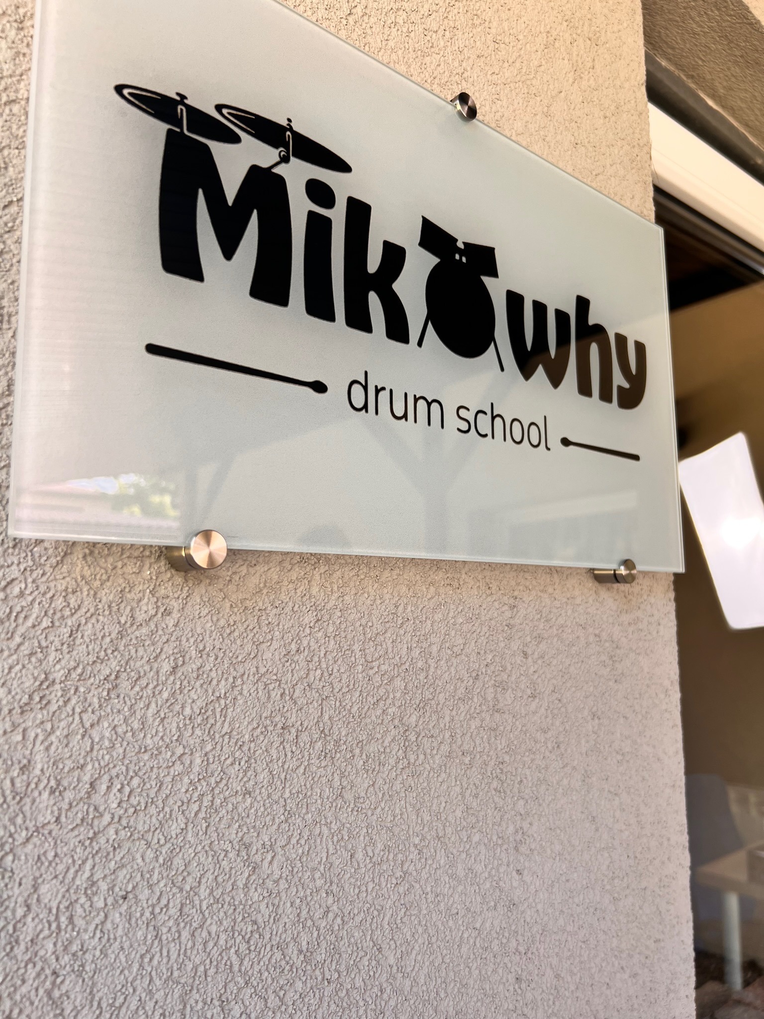 Szyld z nazwą szkoły perkusyjnej 'MikWhy drum school' na szklanej tabliczce przymocowanej do chropowatej ściany zewnętrznej budynku.