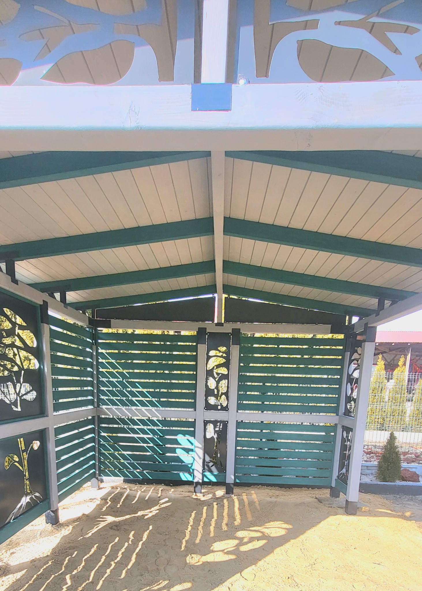 Wnętrze altany z drewnianymi panelami z wyciętymi motywami roślinnymi, piaskowa podłoga oświetlona promieniami słońca padającymi przez szczeliny w panelach.