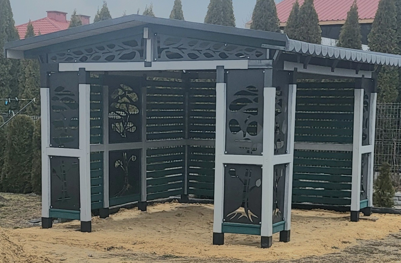 Nowoczesna altana z ażurowymi panelami dekorowanymi motywami drzew, zlokalizowana na posesji z widocznymi tujami i domem w tle.