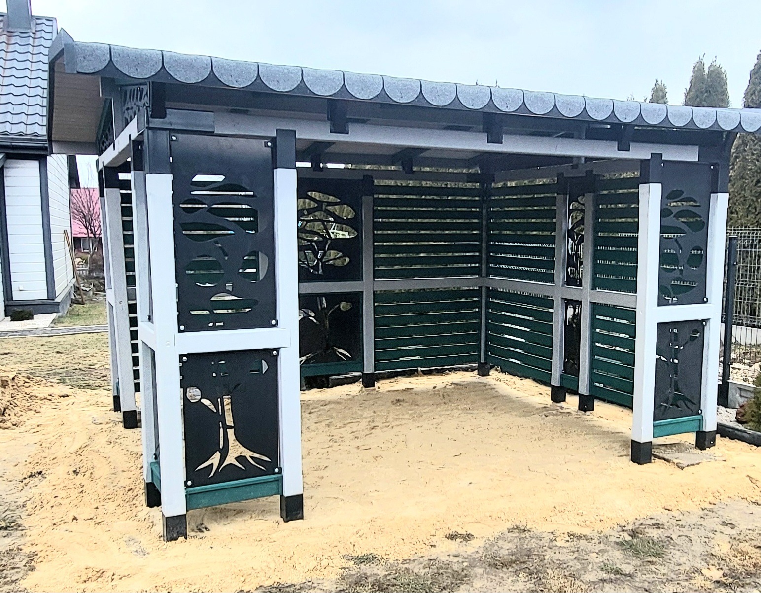 Nowoczesna altana ogrodowa z szarym dachem, białymi ramami i czarnymi panelami dekoracyjnymi z motywem drzewa, otoczona piaskiem.