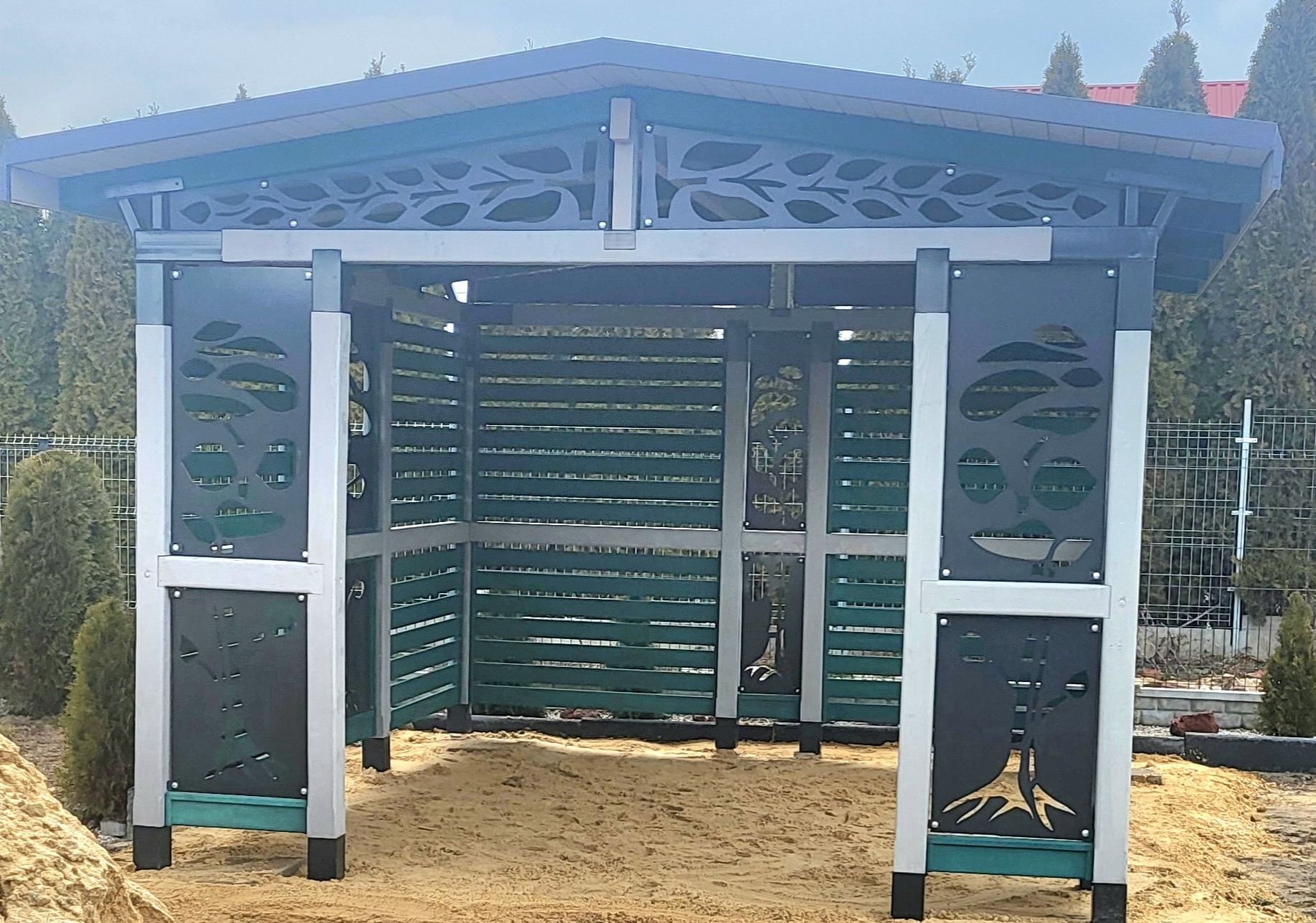 Nowoczesna konstrukcja z ażurowymi panelami z motywem liści, prawdopodobnie altana lub pergola, na piaszczystym podłożu.