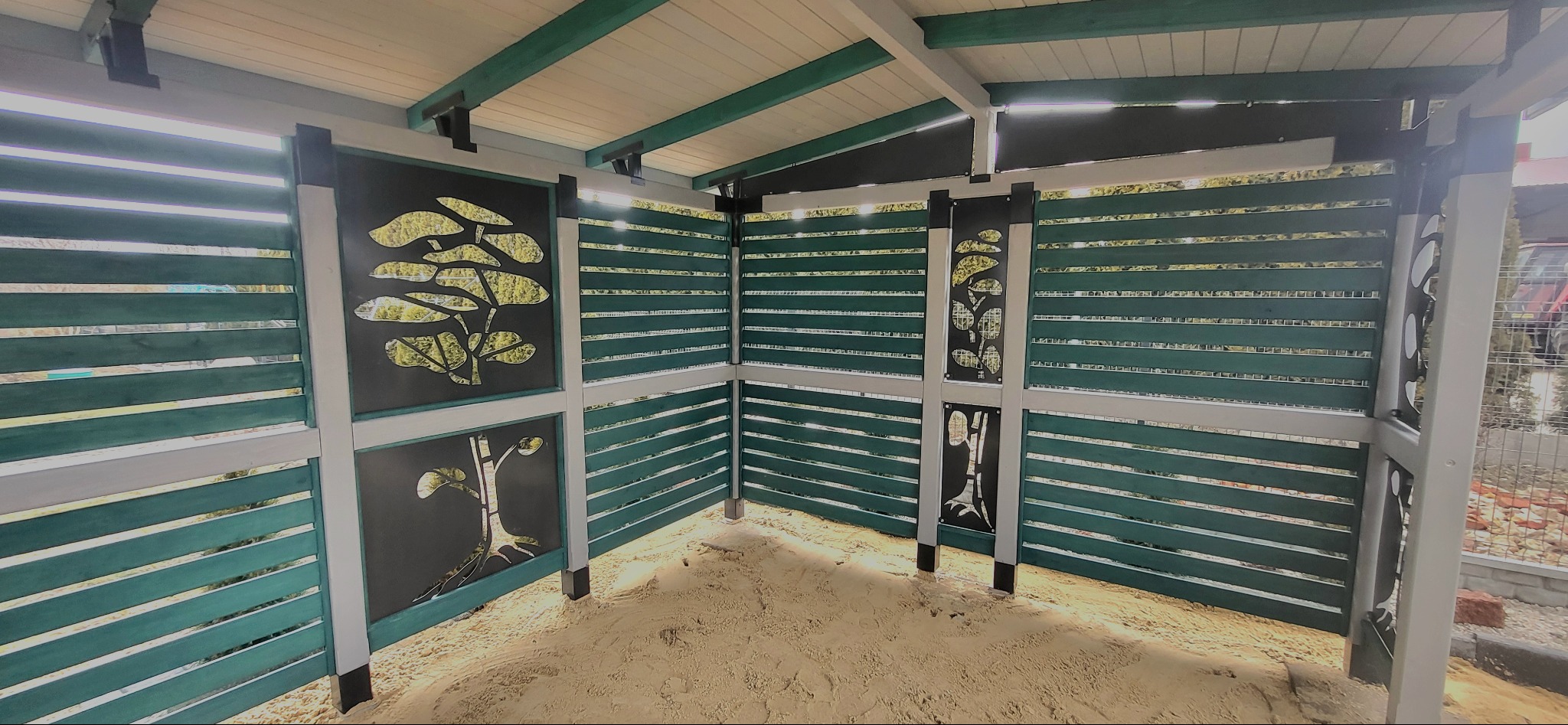 Kącik zabaw dla dzieci z piaskiem, otoczony drewnianą konstrukcją z ażurowymi panelami z motywem roślinnym, pomalowany na zielono i szaro.