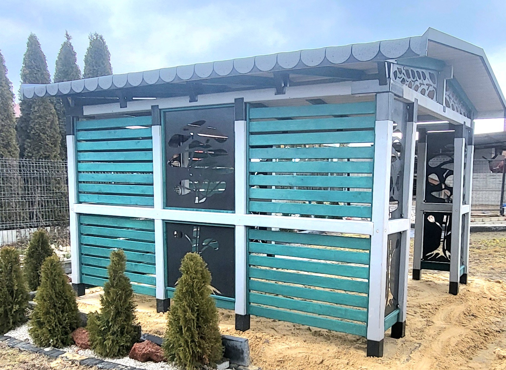 Nowoczesna konstrukcja z drewnianymi panelami w odcieniach turkusu i czarnymi wstawkami z motywem roślinnym, otoczona małymi tujami i piaskiem.