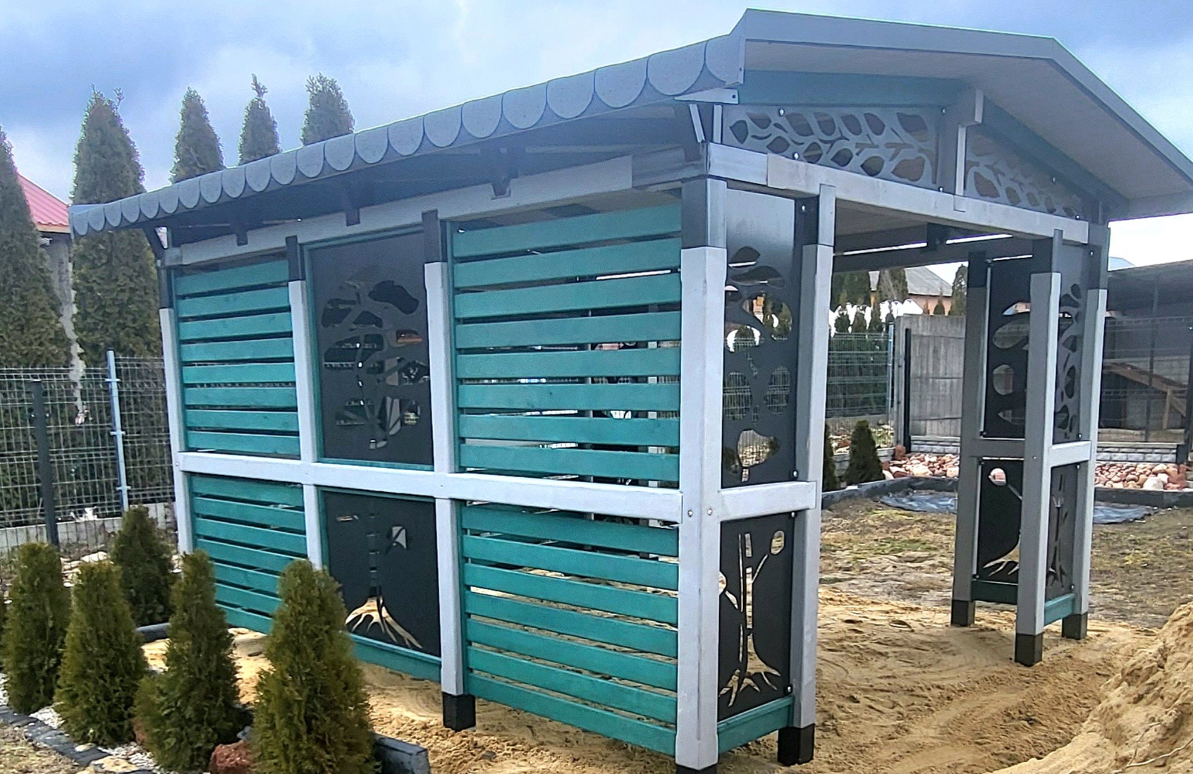 Nowoczesna altana z turkusowymi panelami i dekoracyjnymi wycięciami w kształcie drzew, otoczona młodymi tujami na piaszczystym podłożu.