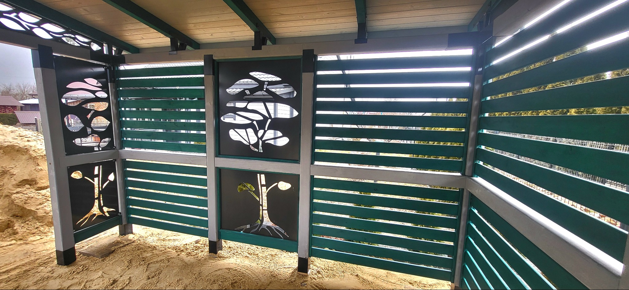 Nowoczesna altana z zielonymi, poziomymi panelami i dekoracyjnymi, wycinanymi laserowo motywami roślinnymi w czarnych panelach, wypełniona piaskiem.