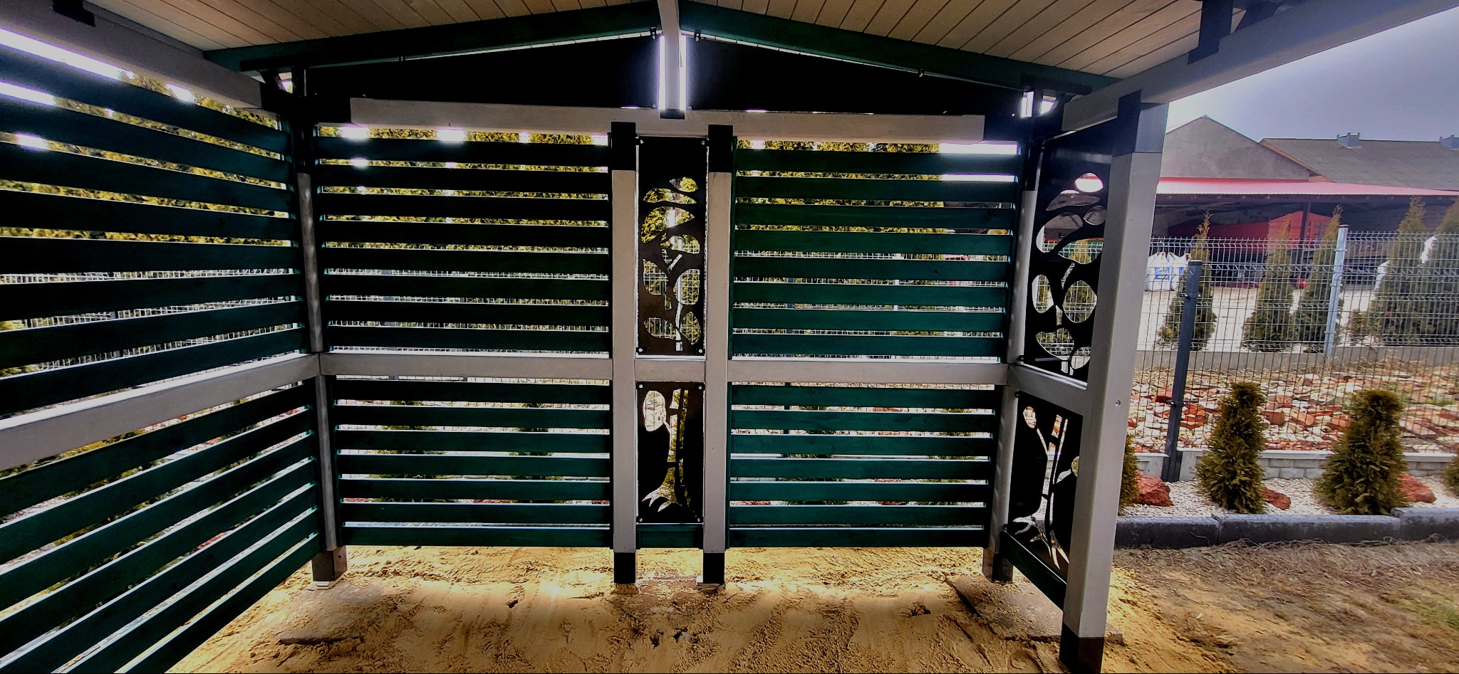 Nowoczesna altana z poziomymi, zielonymi panelami i dekoracyjnymi, wycinanymi elementami w kolorze czarnym, na piaszczystym podłożu, z widokiem na ogród i budynek w tle.