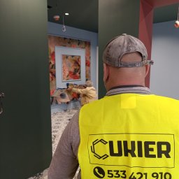 Szymon Cukiernik Centrum Usług - Pracownik w kamizelce z logo firmy maluje ścianę w pomieszczeniu z wzorzystą tapetą i dekoracyjną podłogą. Widok zza pleców, wnętrze w trakcie prac wykończeniowych.