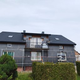 Szymon Cukiernik Centrum Usług - Dom w trakcie ocieplania styropianem, widoczne rusztowania i fragment balkonu z ozdobną balustradą. Elewacja w szarym kolorze, dach pokryty czarną dachówką.
