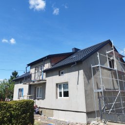 Szymon Cukiernik Centrum Usług - Dom w trakcie tynkowania elewacji, widoczne rusztowania i pracownik przy pracy. Świeży tynk o szarej barwie, błękitne niebo w tle. Ujęcie z dołu, podkreślające wysokość budynku.