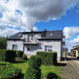 Szymon Cukiernik Centrum Usług - Szary dom jednorodzinny ze stalowymi rusztowaniami w Starowej Górze, częściowo zasłonięty przez zielone krzewy. Widoczne okna, balkon i ciemny dach.