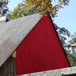 Szymon Cukiernik Centrum Usług - Fragment budynku z szarą, bitumiczną dachówką i intensywnie czerwoną ścianą pokrytą membraną. Widoczne drzewa i fragment komina z wentylatorem. Ujęcie z dołu.