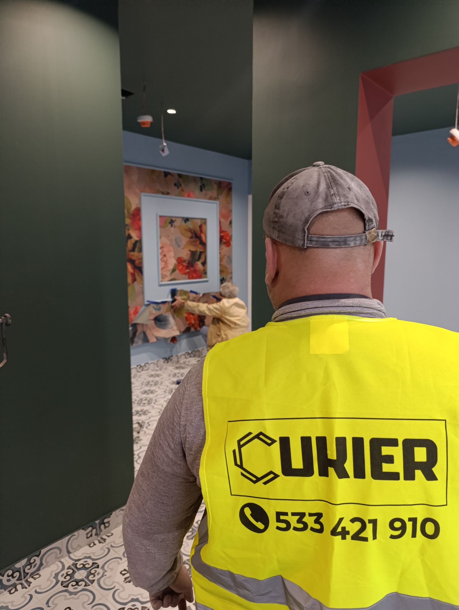 Pracownik w kamizelce z logo firmy maluje ścianę w pomieszczeniu z wzorzystą tapetą i dekoracyjną podłogą. Widok zza pleców, wnętrze w trakcie prac wykończeniowych.