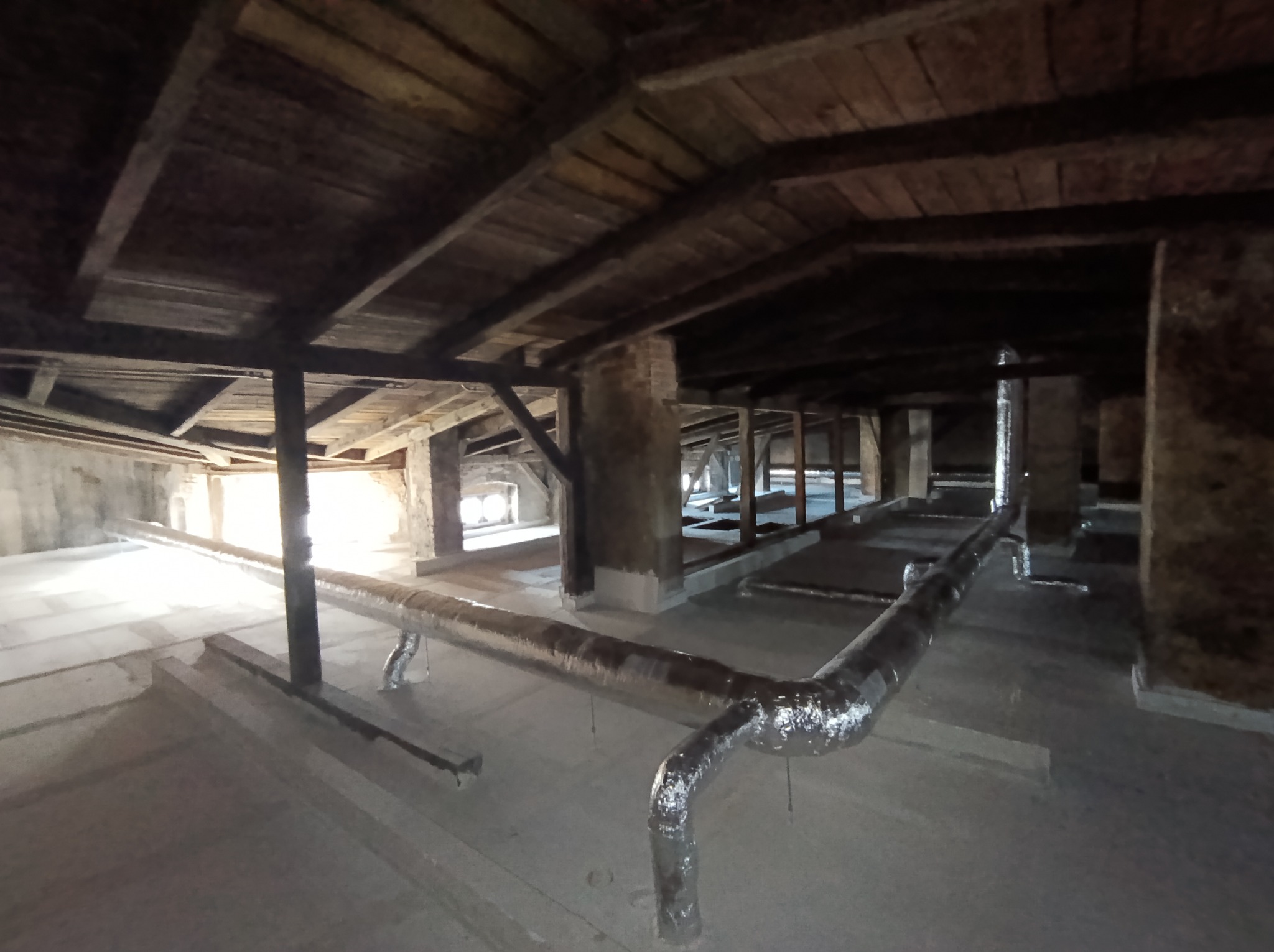 Stare poddasze z drewnianą konstrukcją stropu i rurami instalacji. Widoczne elementy konstrukcyjne i fragmenty instalacji w surowym stanie. Ujęcie z dołu, perspektywa zbieżna.