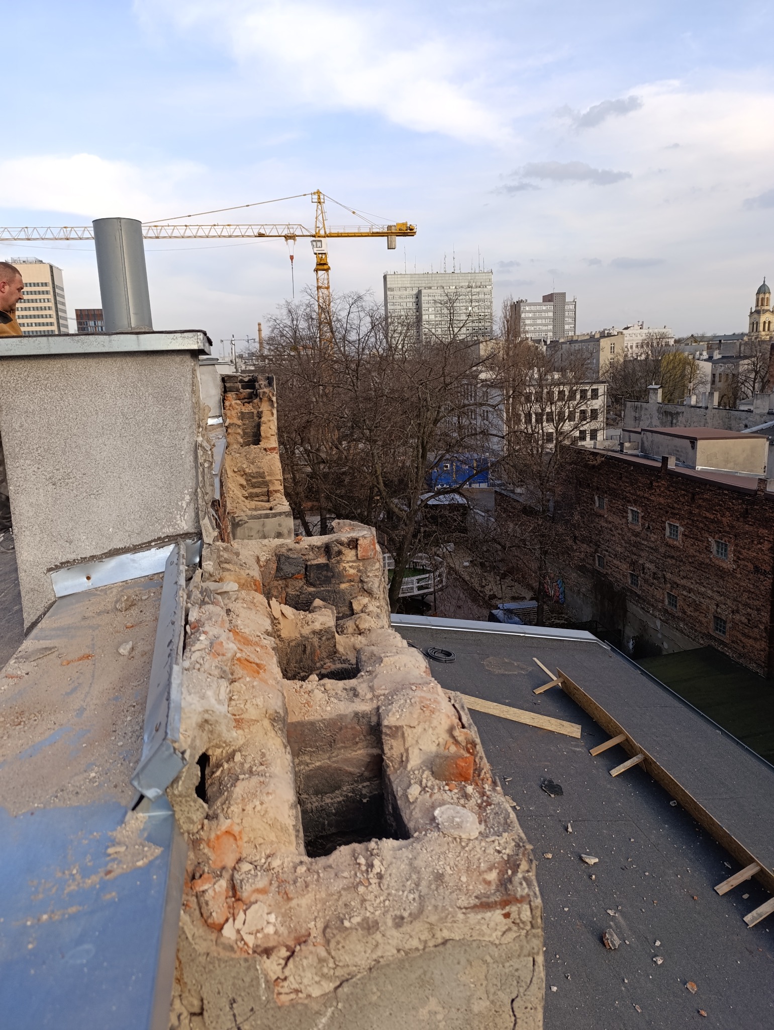 Uszkodzony komin z cegły na dachu budynku w trakcie rozbiórki, z widocznymi elementami konstrukcyjnymi i panoramą miasta w tle. Prace rozbiórkowe w toku.