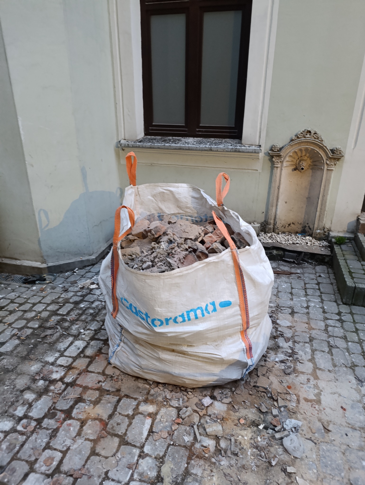 Worek z gruzem po rozbiórce na brukowanym podłożu, obok ściana budynku z oknem i ozdobną fontanną. Pomarańczowe uchwyty worka. Widoczne pozostałości po pracach.