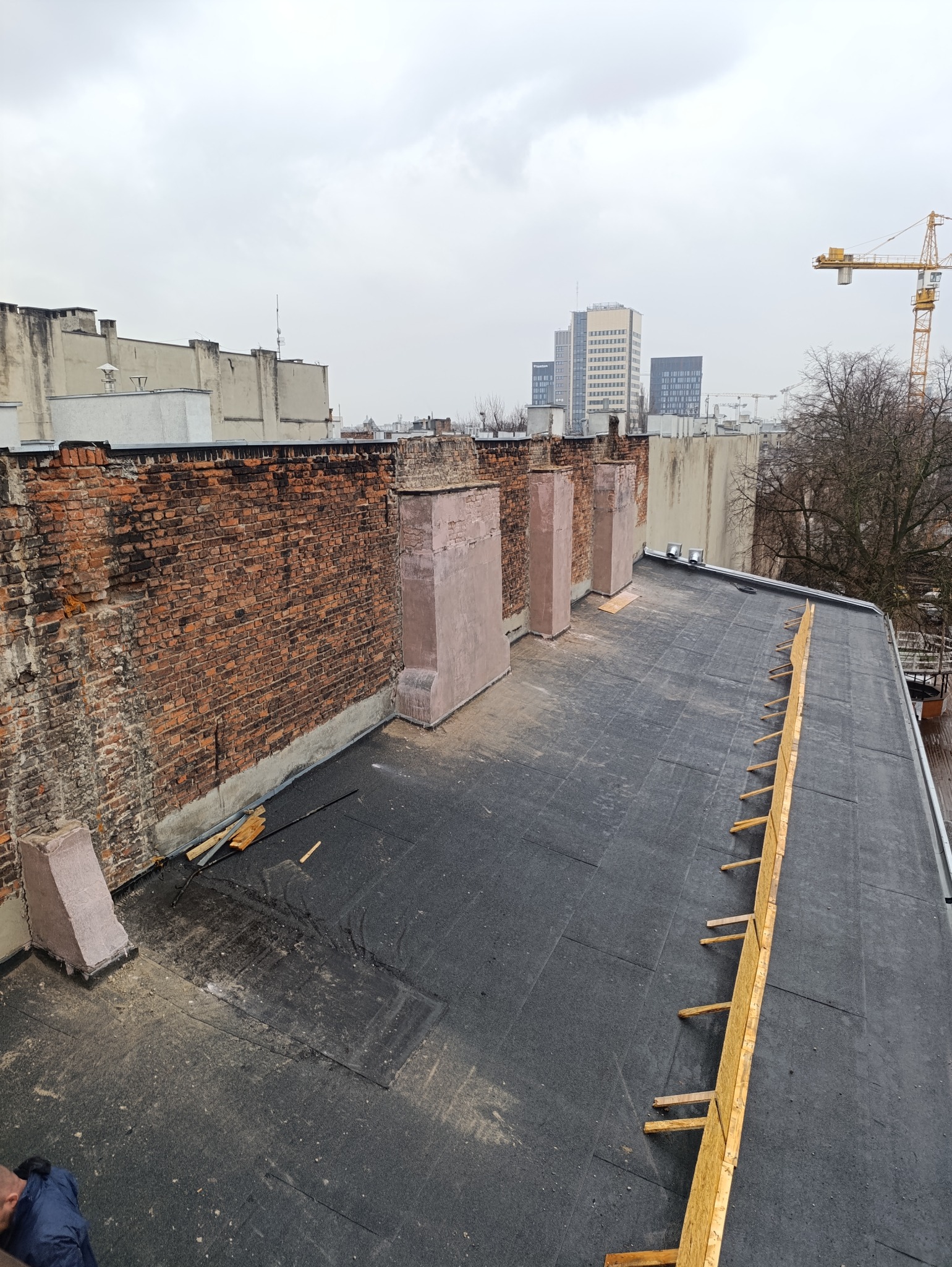 Widok z góry na płaski dach pokryty czarną papą, z fragmentem ceglanej ściany i elementami konstrukcyjnymi. W tle zabudowa miejska i dźwig budowlany.