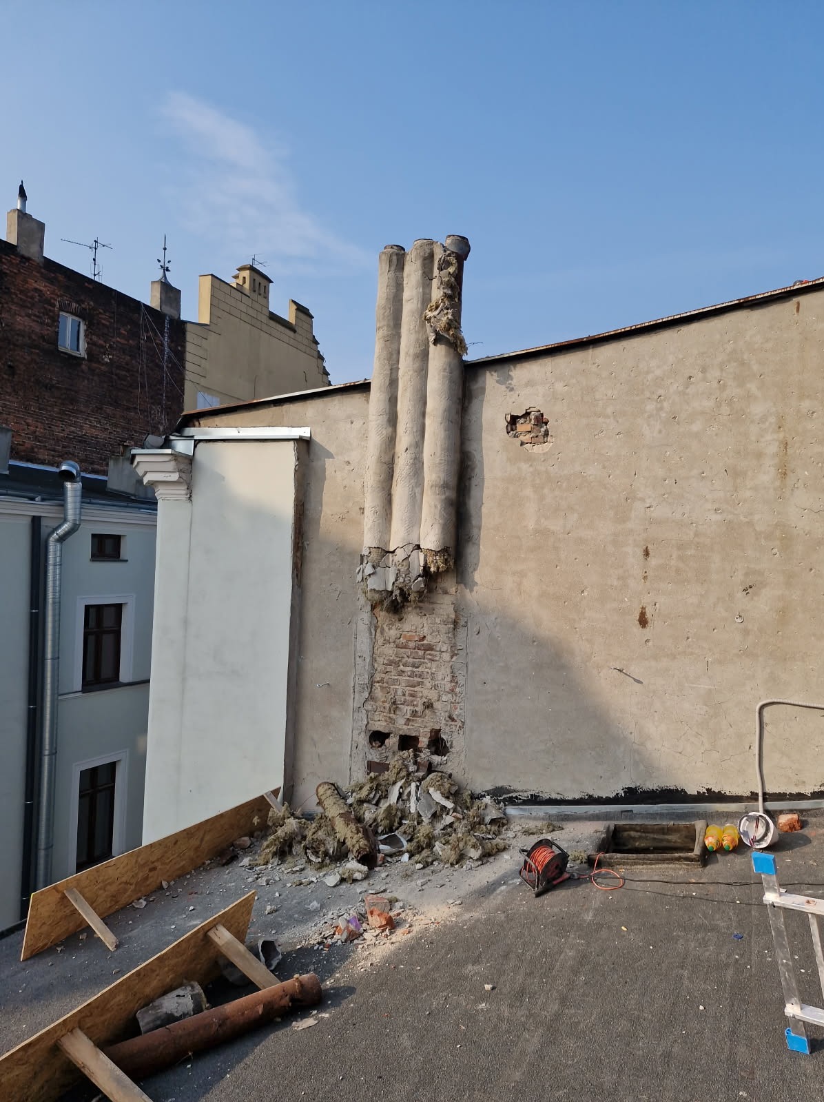 Demontaż komina na dachu budynku. Widoczne pozostałości po kominie, gruz, narzędzia i fragmenty konstrukcji. Prace rozbiórkowe w toku, z bliska, w słoneczny dzień.
