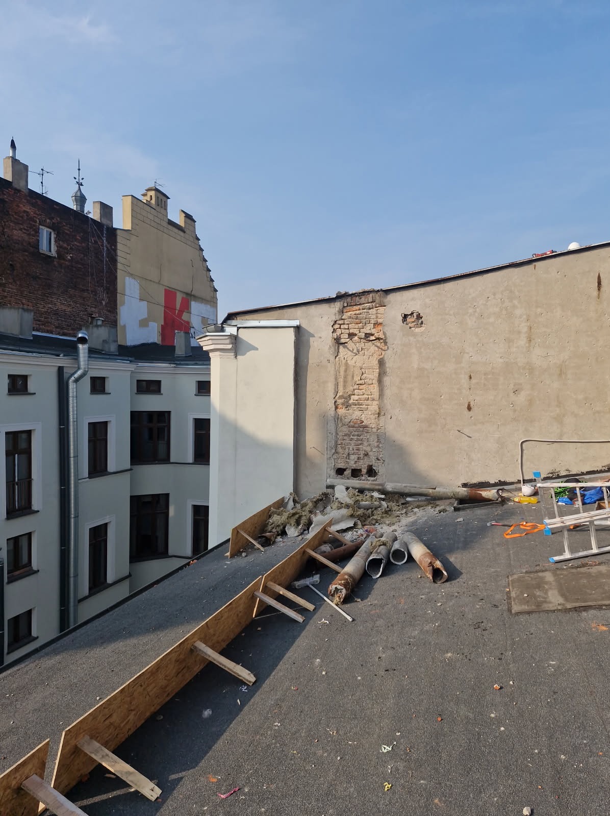 Demontaż komina na dachu budynku. Widoczne fragmenty konstrukcji, rury, izolacja i drabina. W tle zabudowa miejska z budynkami o zróżnicowanej architekturze.