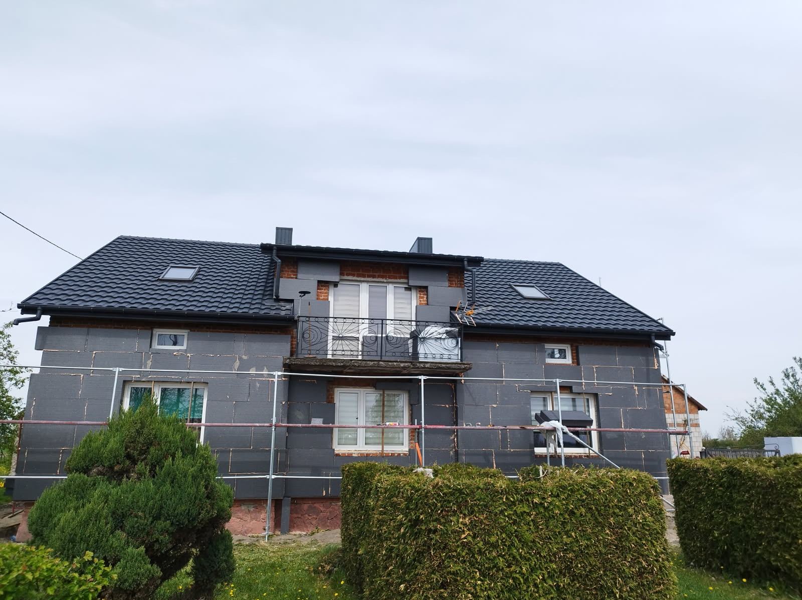 Dom w trakcie ocieplania styropianem, widoczne rusztowania i fragment balkonu z ozdobną balustradą. Elewacja w szarym kolorze, dach pokryty czarną dachówką.