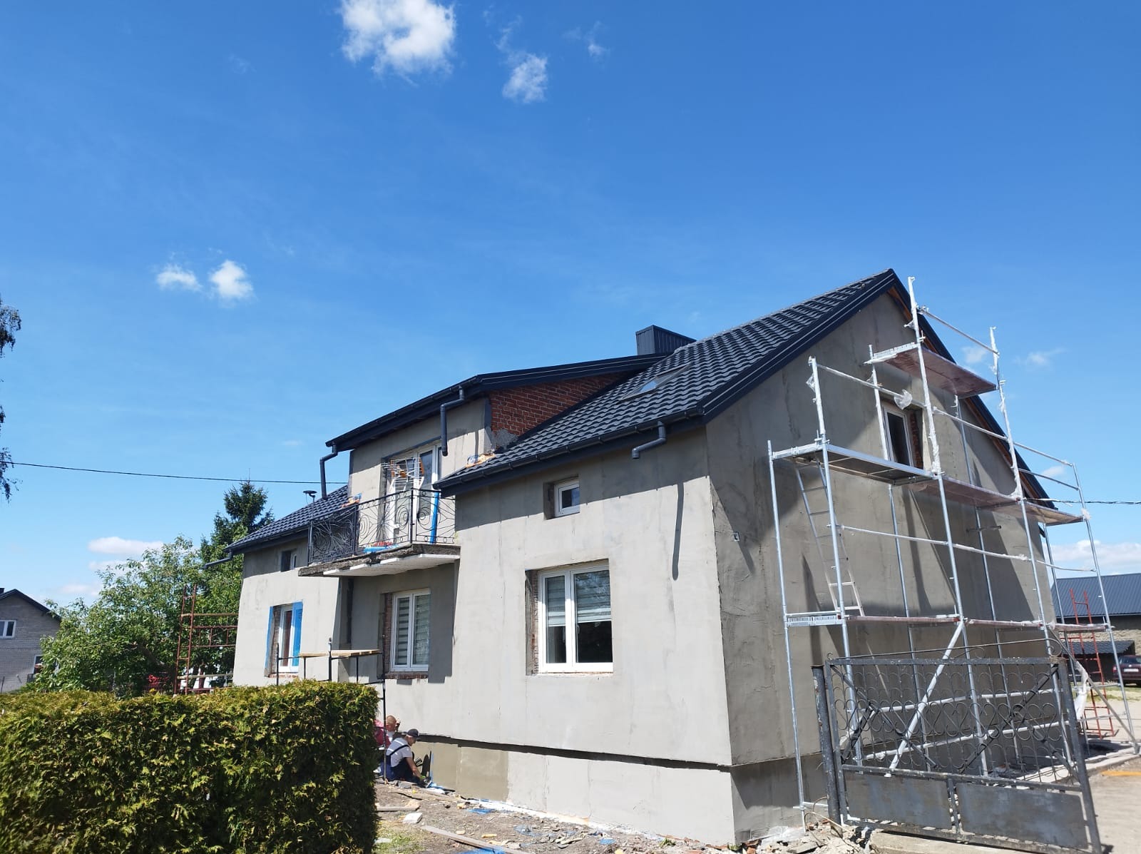 Dom w trakcie tynkowania elewacji, widoczne rusztowania i pracownik przy pracy. Świeży tynk o szarej barwie, błękitne niebo w tle. Ujęcie z dołu, podkreślające wysokość budynku.