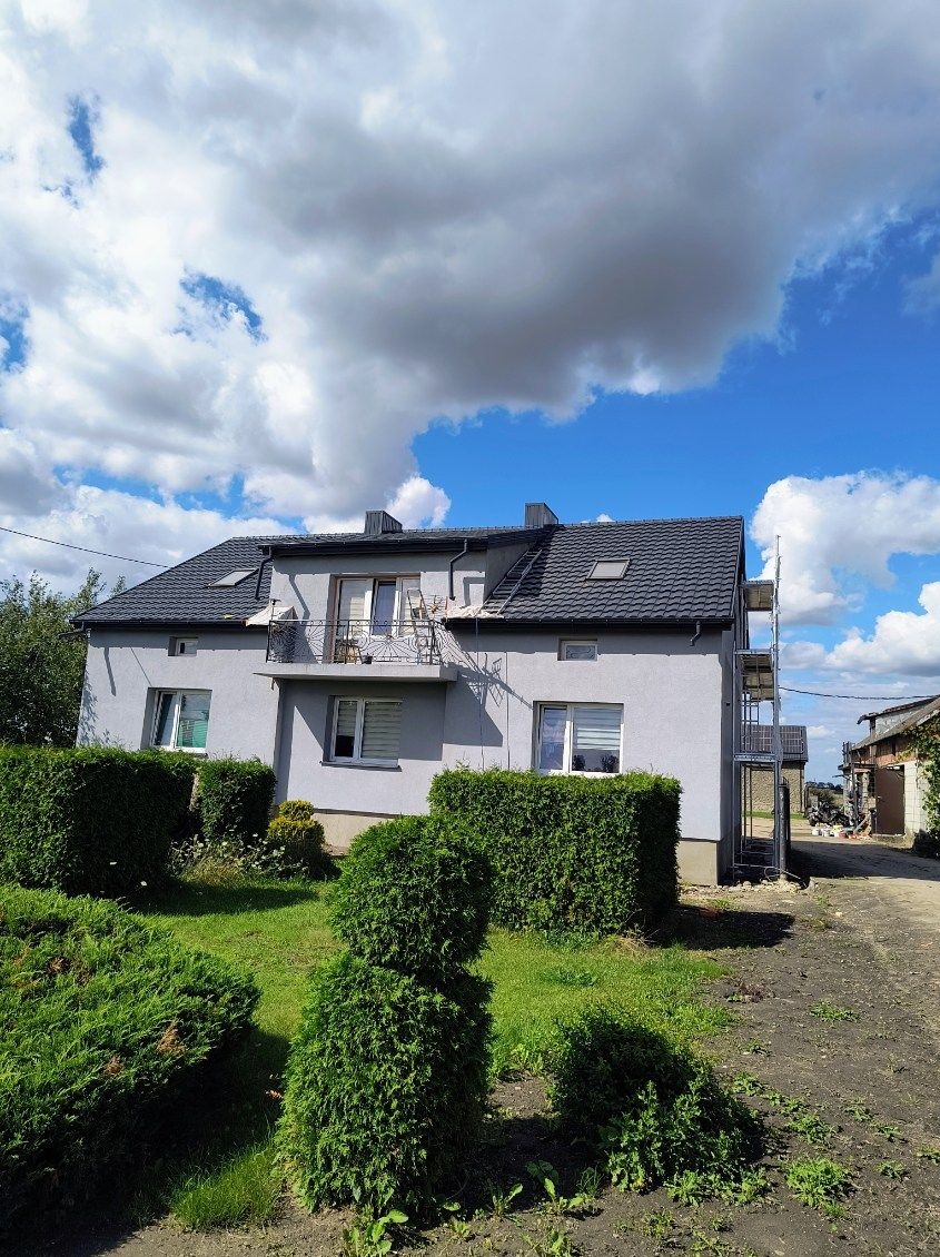 Szary dom jednorodzinny ze stalowymi rusztowaniami w Starowej Górze, częściowo zasłonięty przez zielone krzewy. Widoczne okna, balkon i ciemny dach.