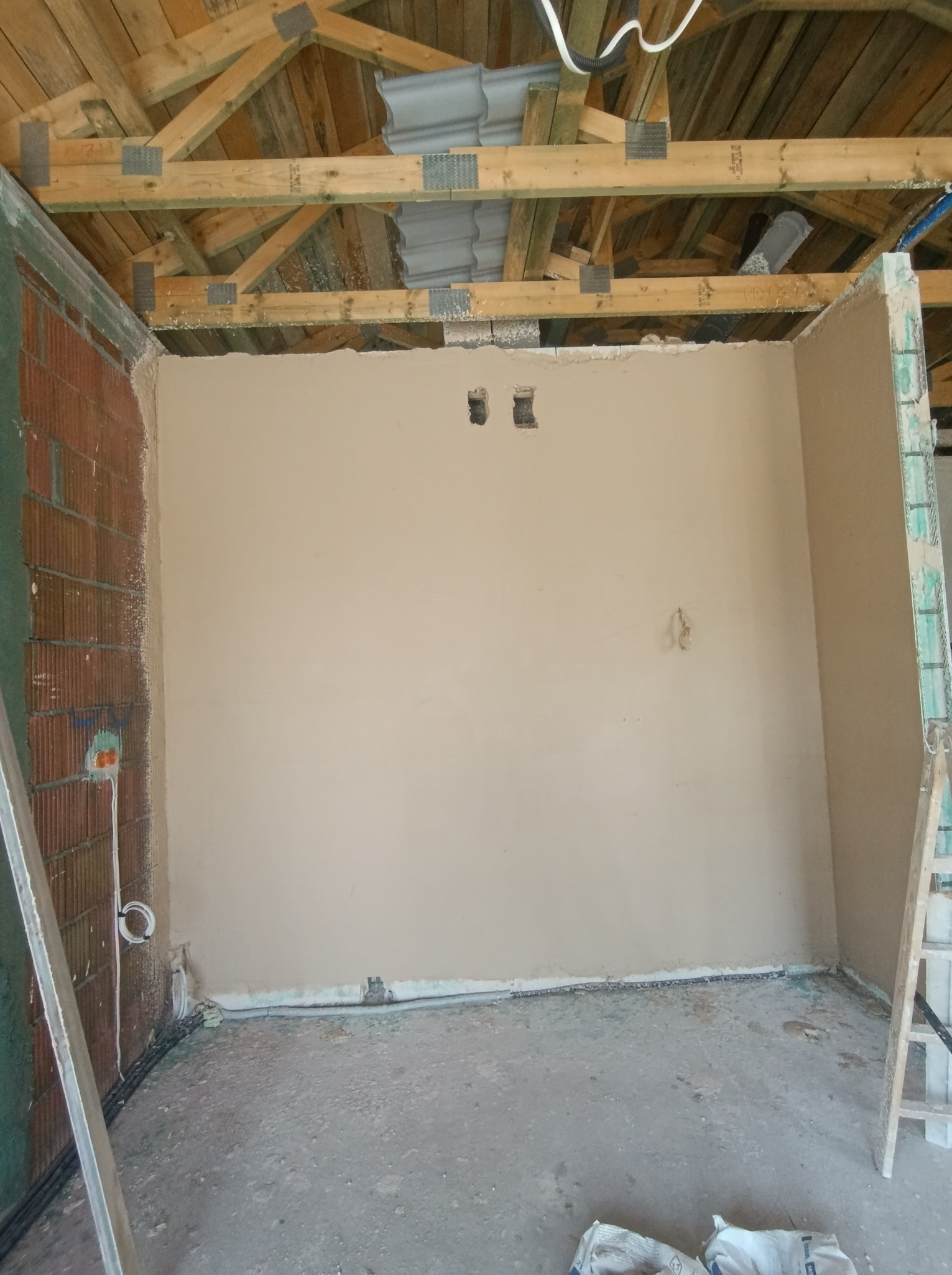 Wnętrze w trakcie tynkowania: ściana pokryta jasnym tynkiem, widoczne przewody elektryczne, cegła, drewniana konstrukcja dachu i drabina. Etap prac wykończeniowych.