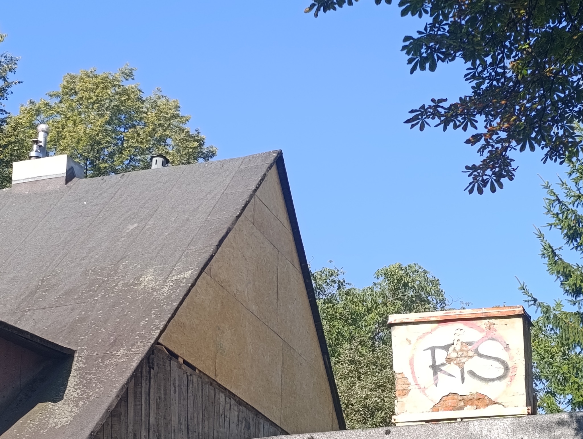Fragment dachu z kominem i ścianą szczytową obłożoną płytami OSB, na tle błękitnego nieba i zieleni drzew. Widoczny element z graffiti.