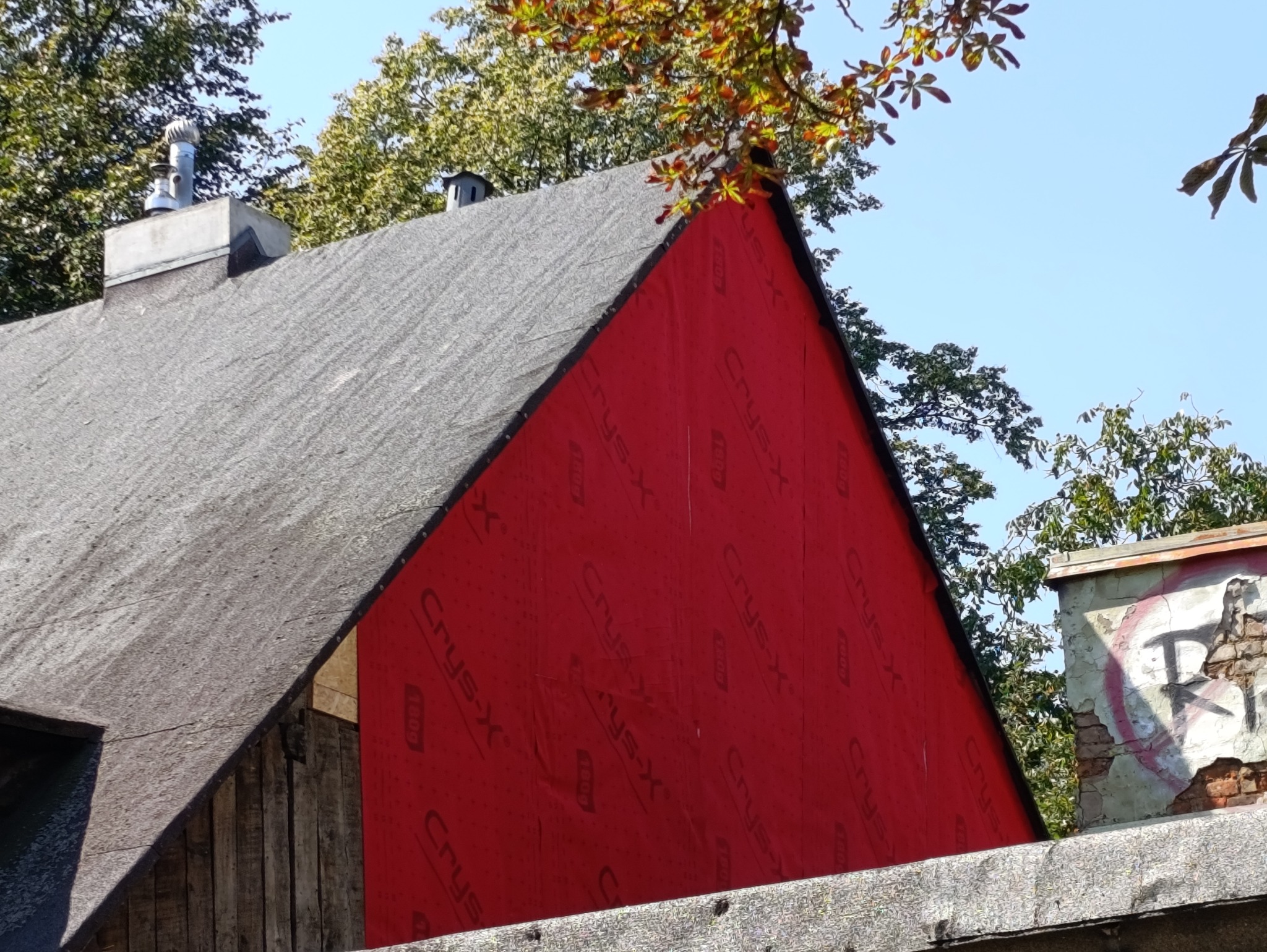Fragment budynku z szarą, bitumiczną dachówką i intensywnie czerwoną ścianą pokrytą membraną. Widoczne drzewa i fragment komina z wentylatorem. Ujęcie z dołu.