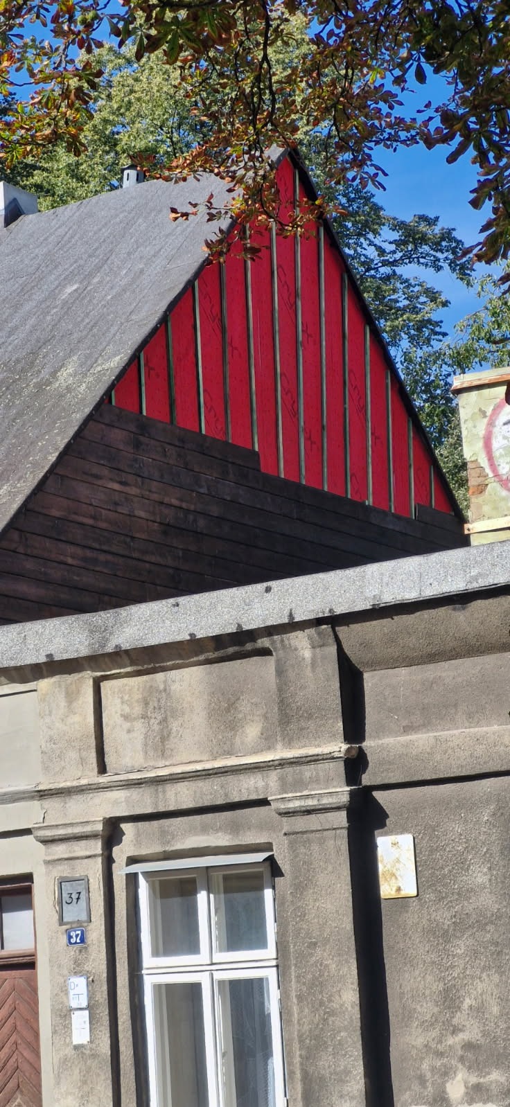 Fragment elewacji budynku z widoczną czerwoną, pionową okładziną i szarą, drewnianą boazerią. Widoczne okno i numer budynku. U góry kadru liście drzew.
