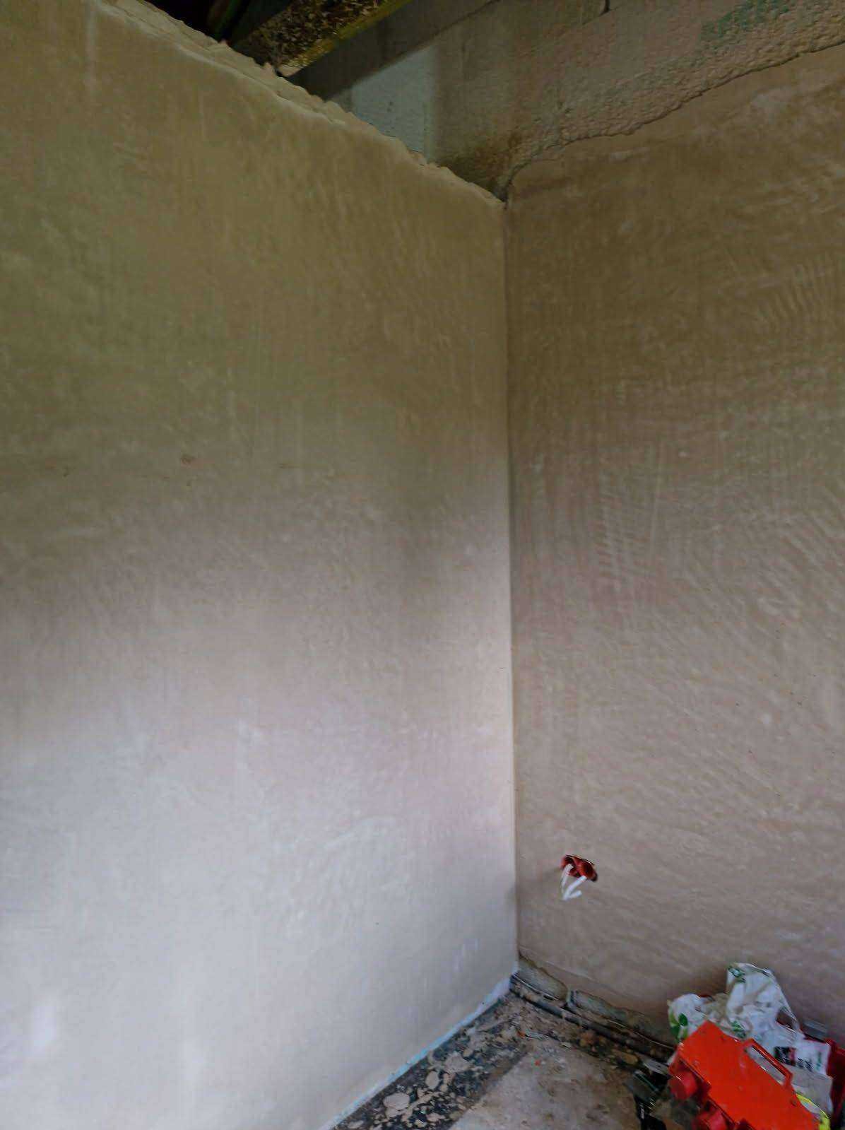 Świeżo nałożony tynk maszynowy na ścianach wewnętrznych w rogu pomieszczenia, widoczne ślady po narzędziach i instalacja elektryczna. Na podłodze resztki materiałów budowlanych.