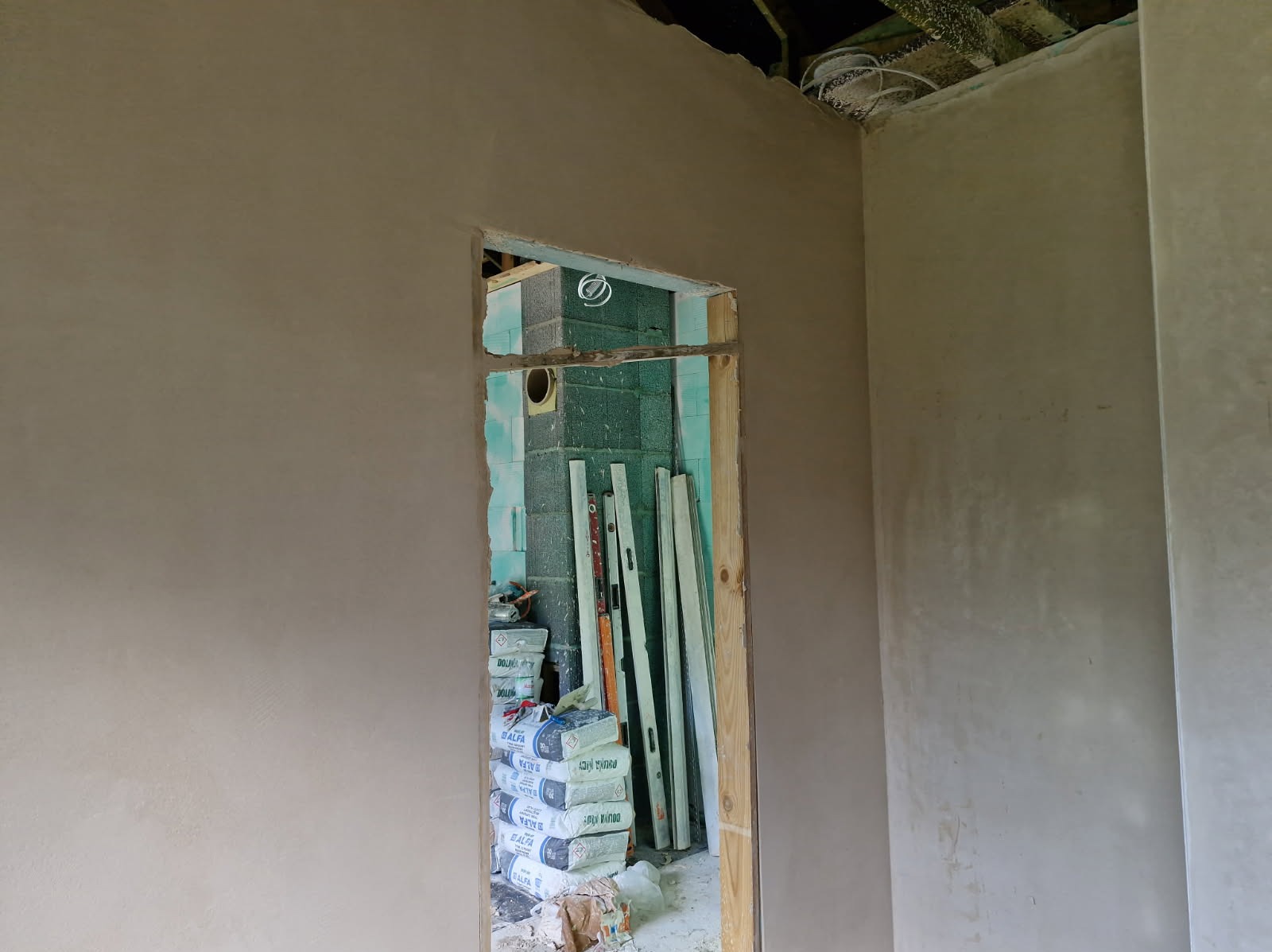 Świeżo położone tynki maszynowe na ścianach wewnątrz budynku, widoczne worki z zaprawą i poziomice w przejściu. Surowy stan wykończenia.