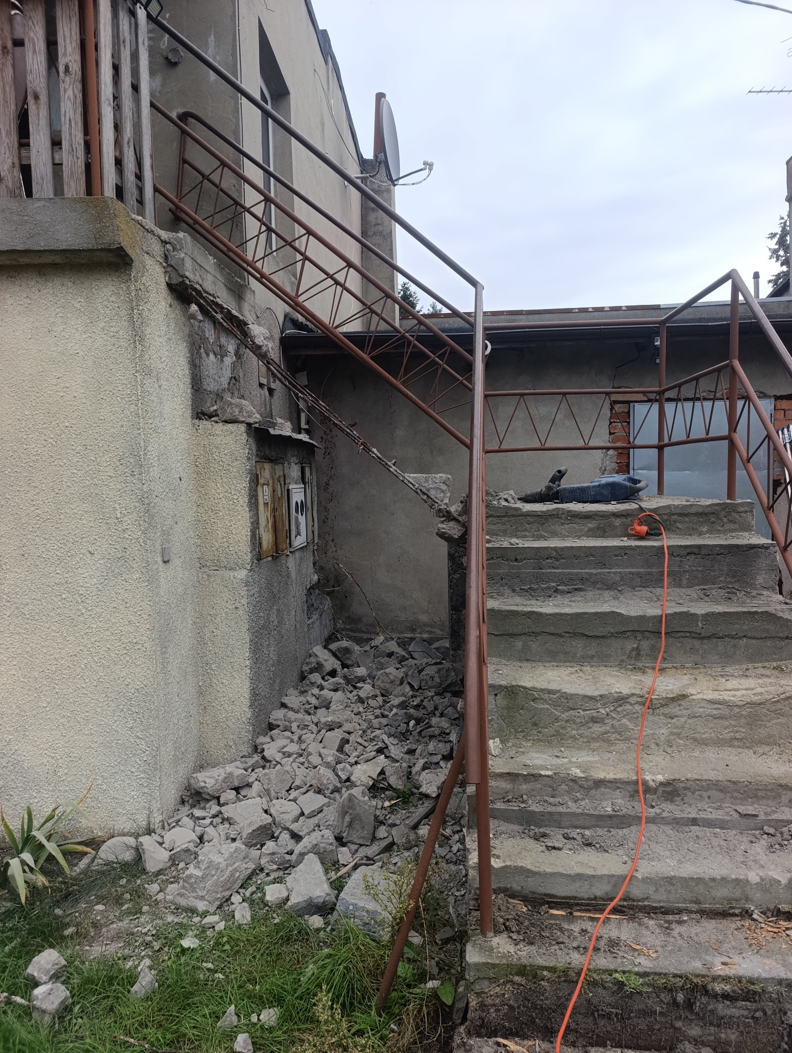 Uszkodzone schody zewnętrzne z gruzem i narzędziami, widoczne ślady rozbiórki. Metalowa balustrada, popękany tynk na ścianie budynku. Prace remontowe w toku.
