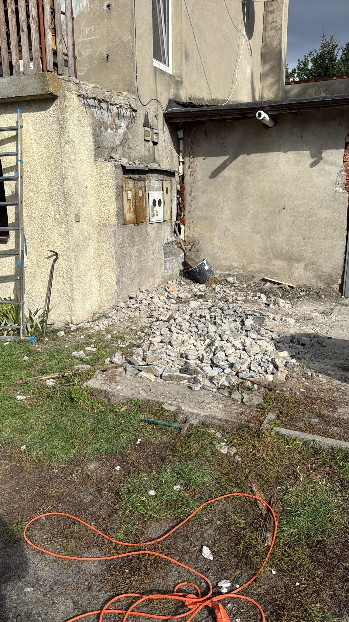 Fragment ściany budynku w trakcie rozbiórki, z widocznymi gruzami, narzędziami i kablem elektrycznym na trawie. Widoczne uszkodzenia tynku i odsłonięta instalacja.