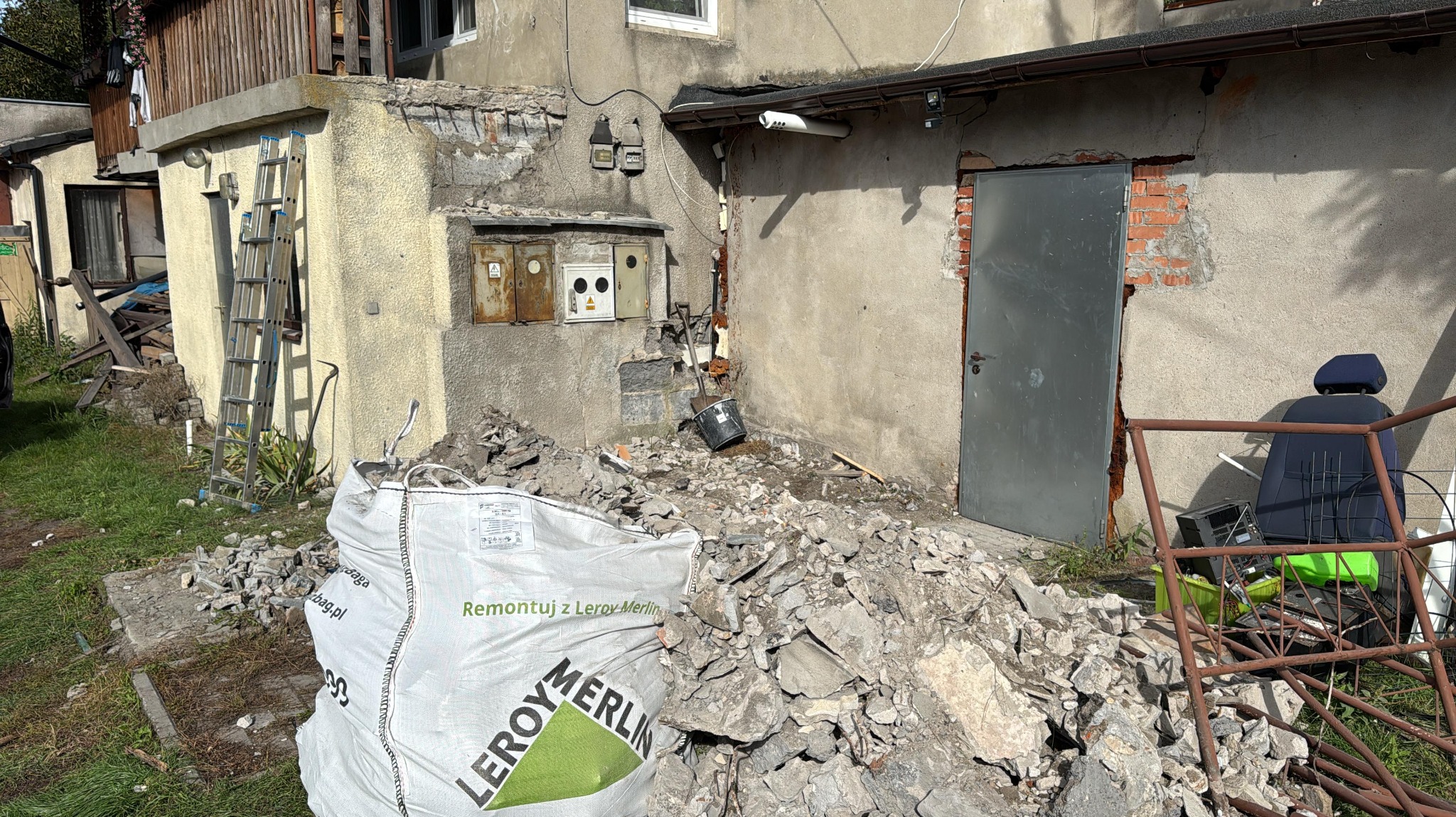 Stary budynek w trakcie rozbiórki, gruz w worku Leroy Merlin, widoczne instalacje elektryczne i metalowe drzwi. Prace rozbiórkowe w toku, bałagan na posesji.
