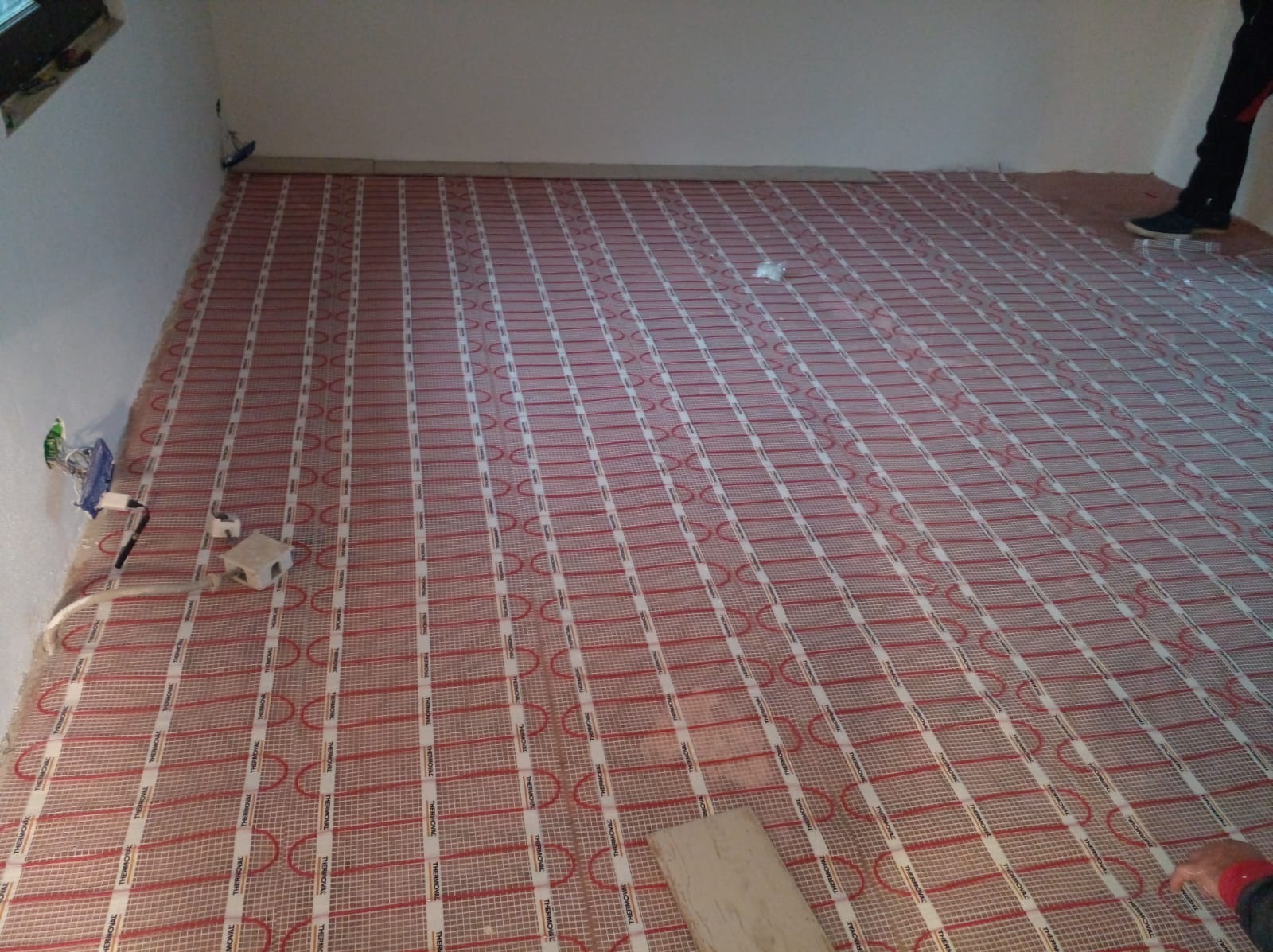 Rozłożona mata grzewcza THERMOVAL na podłodze w pomieszczeniu, przygotowana do zalania wylewką. Widoczne kable i puszka elektryczna na ścianie. Fragment nogi w prawym górnym rogu.