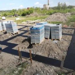 Zalane fundamenty pod dom w kształcie litery L, z widocznymi paletami z bloczkami betonowymi, zbrojeniami i deskowaniem, w tle krajobraz z budynkiem przemysłowym.