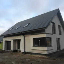 Surowy stan domu jednorodzinnego z ciemną dachówką, trzema oknami dachowymi i dużym oknem narożnym, widoczne rynny i obróbki blacharskie, elewacja w trakcie wykończenia.