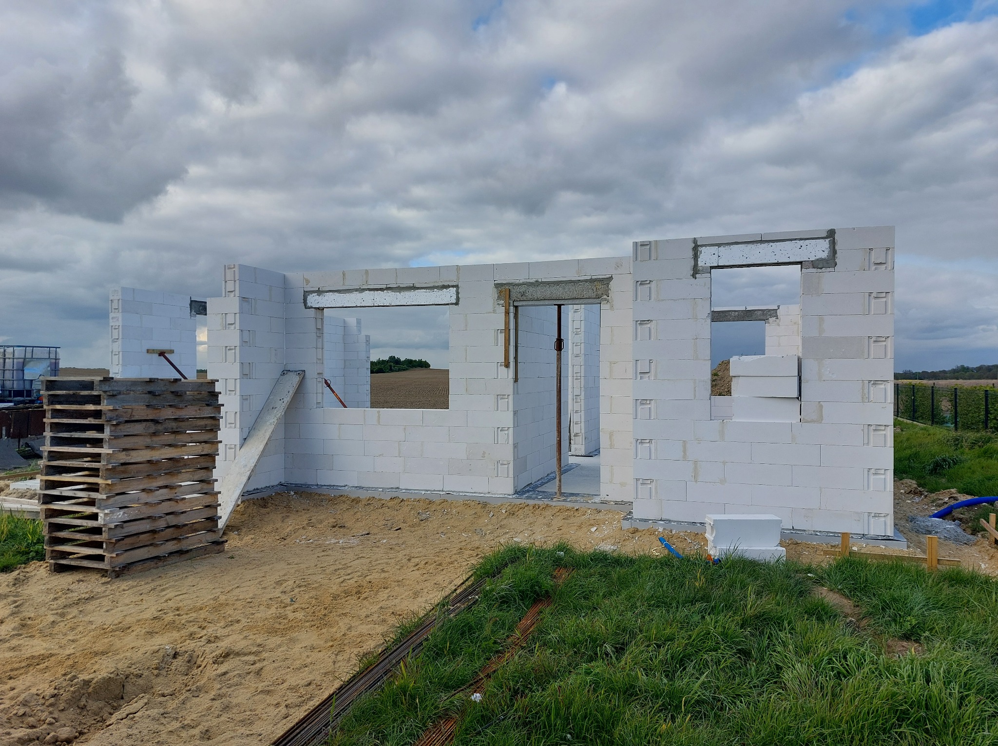 Wznoszone ściany parterowego domu z białych bloczków, widoczne otwory okienne i drzwiowe, na pierwszym planie paleta z bloczkami i zbrojenie, w tle pochmurne niebo.