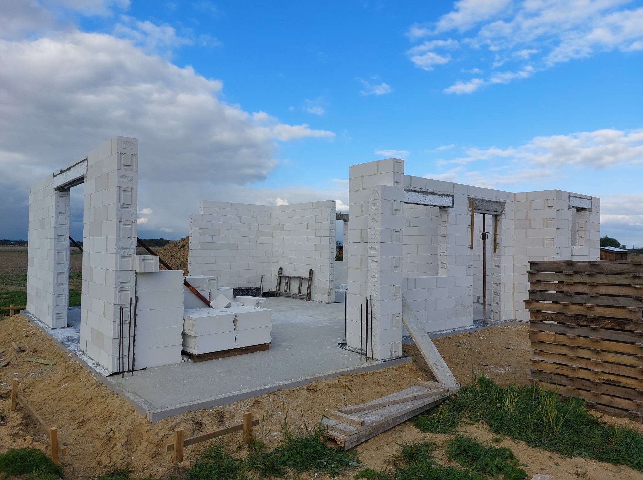 Wznoszenie ścian parterowego domu z betonu komórkowego Ytong, widoczne zbrojenia pionowe, paleta z bloczkami oraz drewniana drabina wewnątrz budynku, fundament wylany.