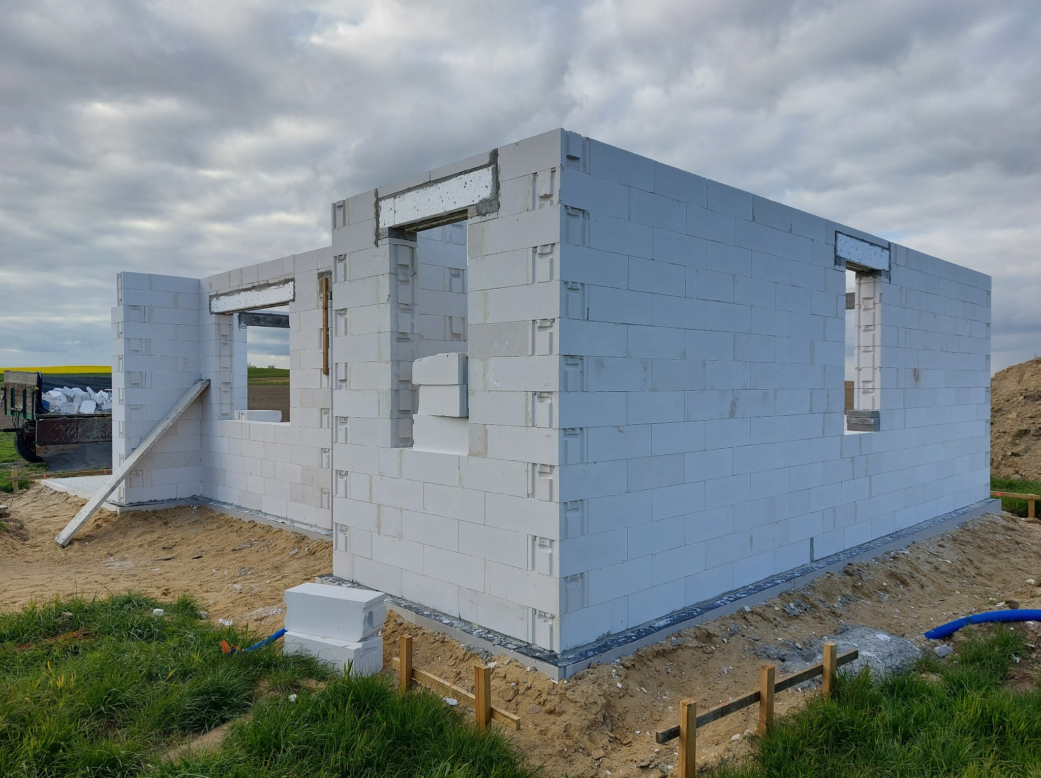 Budowa domu murowanego z białych bloczków, widoczne otwory okienne i drzwiowe z nadprożami, częściowo wykończone fundamenty, teren budowy z trawą i piaskiem, pochmurne niebo.