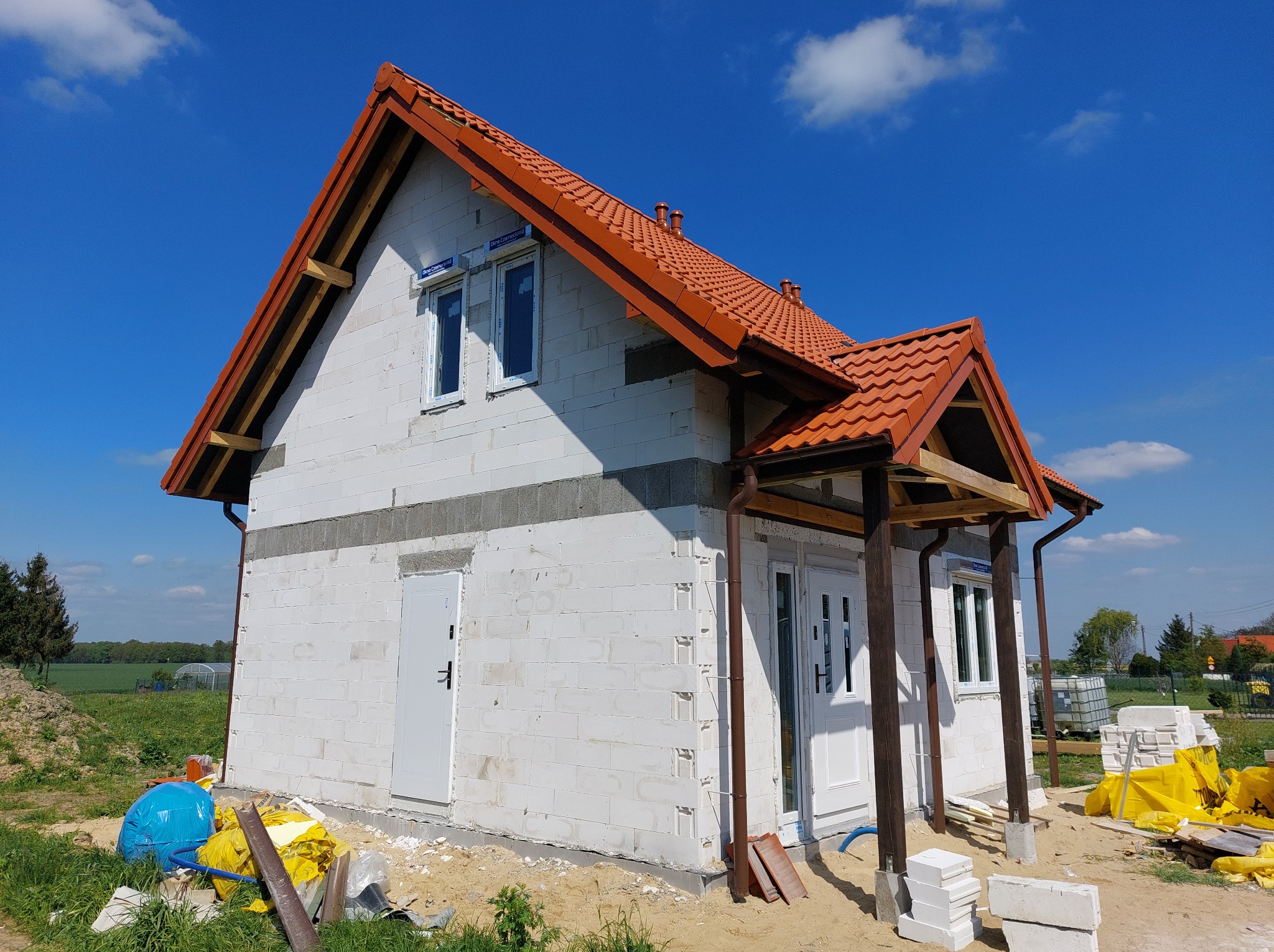 Surowy stan domu z białych bloczków z czerwoną dachówką, widoczne okna z roletami, niezadaszone wejście z drewnianymi słupami, teren budowy z materiałami budowlanymi w tle.
